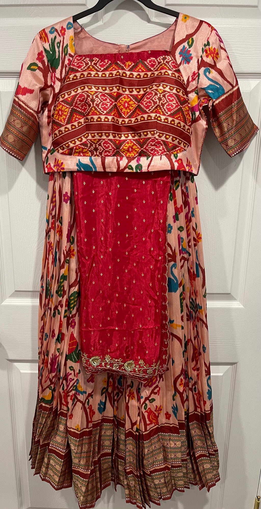 A traditional Indian salwar suit set hanging on a black hanger against a white paneled door. The set includes a pink and red printed kurta with geometric patterns, a matching red embroidered skirt, and a red embroidered blouse panel.