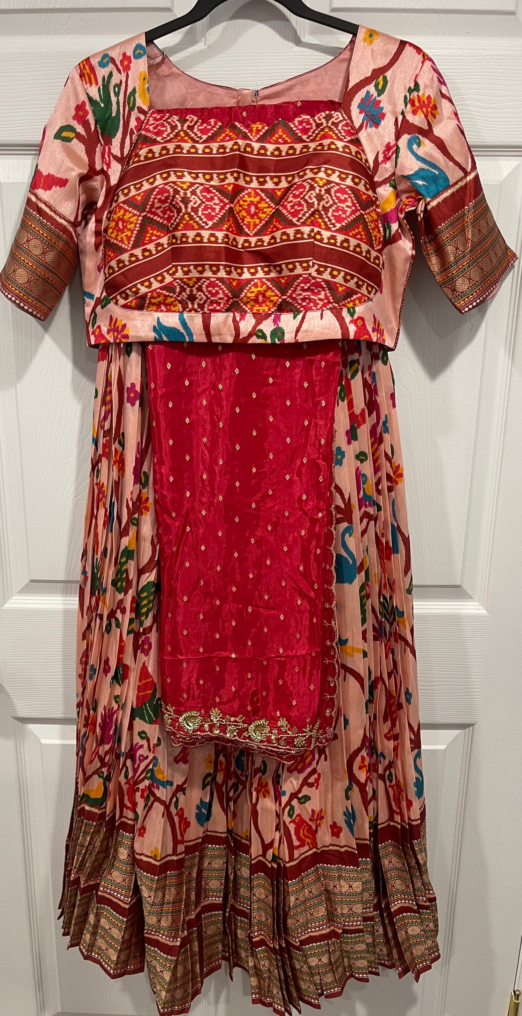 A traditional Indian salwar suit set hanging on a black hanger against a white paneled door. The set includes a pink and red printed kurta with geometric patterns, a matching red embroidered skirt, and a red embroidered blouse panel.